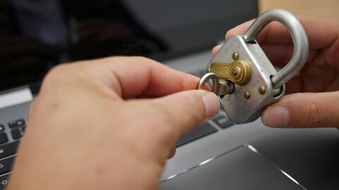 Zwei Hände, die ein Vorhängeschloss schließen, im Hintergrund ein Laptop.