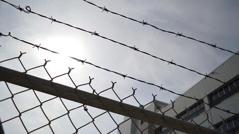 Stacheldrahtzaun, im Hintergrund ist ein Stück eines Betonbaus zu sehen.